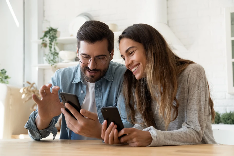Couple souriant regardant ensemble leurs smartphones dans un intérieur moderne lumineux avec étagères blanches et plantes vertes