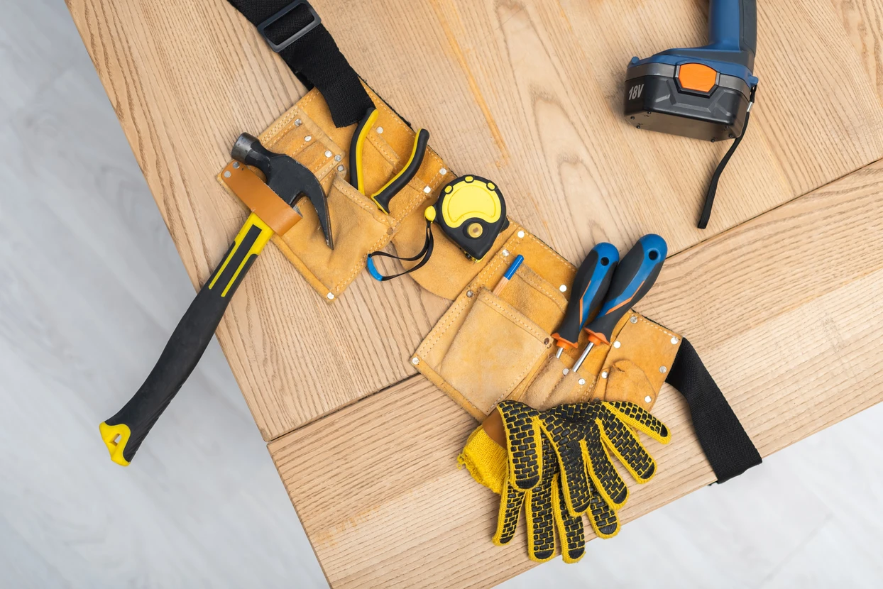 Intérieur, plan de travail en bois avec ceinture porte-outils en cuir beige contenant marteau à manche noir et jaune, tournevis à manche bleu, mètre ruban jaune et noir, gants de protection jaunes antidérapants
