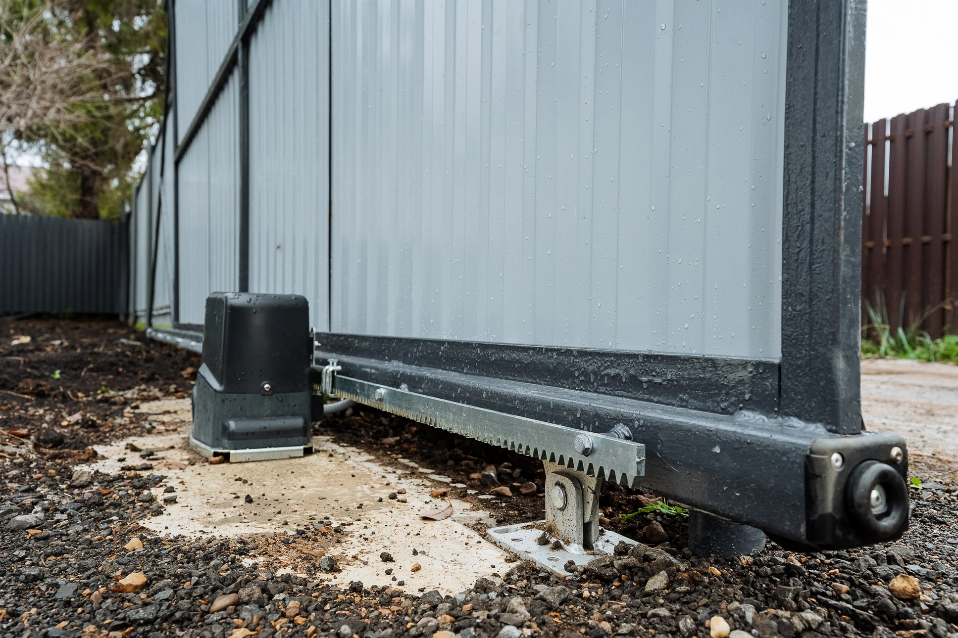 Photographie professionnelle en plan moyen montrant un portail en fer forgé ou aluminium moderne d'une résidence, avec ses piliers en maçonnerie. Le portail est partiellement ou complètement ouvert, révélant le système de motorisation visible : motoréducteur, bras de commande, ou mécanisme d'entraînement installé sur l'un des piliers ou au-dessus du portail. Un électricien professionnel en tenue de travail est présent, effectuant une vérification ou une intervention finale sur le système. Le décor est résidentiel, lumineux, authentique, sans effets numériques.